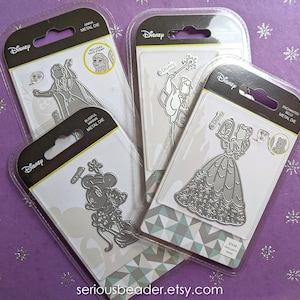 May include: Four Disney metal dies in clear plastic packaging. The dies feature characters Anna, Cinderella, Minnie Mouse, and a fairy. The packaging includes the Disney logo and the words "METAL DIE". The background is a purple surface with silver snowflake accents.