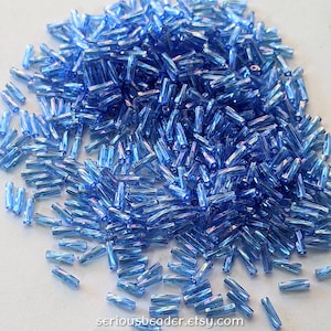 May include: A pile of iridescent blue glass beads. The beads are cylindrical with a faceted surface, catching the light and creating a shimmering effect. The beads are scattered, suggesting they are for jewelry making or crafts.