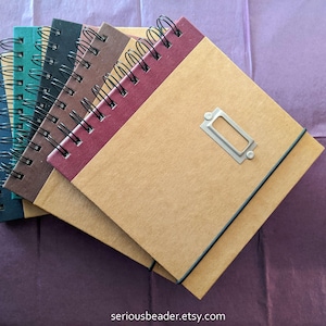 May include: A stack of five brown, burgundy, and green spiral bound notebooks with a metal label holder on the top notebook. The notebooks are on a purple surface.
