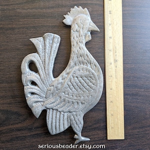 May include: A silver-colored metal rooster wall hanging. The rooster is approximately 10 inches tall and has detailed feather and comb designs. The Etsy shop name "seriousbeader.etsy.com" is printed at the bottom.