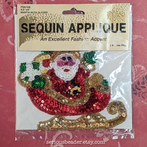 May include: A sequin applique of Santa Claus in a sleigh, packaged in a clear plastic bag with a header card labeled FSA128 4.5"x4" SANTA WITH SLEIGH. The applique features red, white, green, and gold sequins depicting Santa with a gift sack.