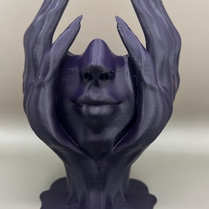May include: A dark purple 3D printed sculpture of a face with hands covering the eyes. The sculpture is abstract and has a smooth, matte finish.