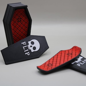 May include: Black coffin-shaped box with a red diamond-patterned interior, and two miniature skateboards. One skateboard is red with a black diamond pattern, and the other is black with the word "FLIP" and a skull.
