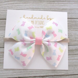 K&ouml;nnte beinhalten: Eine wei&szlig;e Schleife mit einem pastellfarbenen Hasenmuster, gesichert mit einer rosa Mitte und einem beigen Haarband. Die Schleife befindet sich auf einer wei&szlig;en Karte mit goldenem Text "handmade by MB DESIGNS".