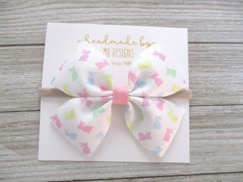 K&ouml;nnte beinhalten: Eine wei&szlig;e Schleife mit einem pastellfarbenen Hasenmuster, gesichert mit einer rosa Mitte und einem beigen Haarband. Die Schleife befindet sich auf einer wei&szlig;en Karte mit goldenem Text "handmade by MB DESIGNS".