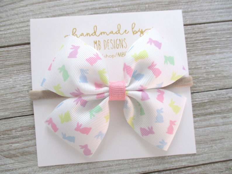K&ouml;nnte beinhalten: Wei&szlig;es Schleifenband mit pastellfarbenem Hasenmuster, rosa Mitte und einem neutralfarbenen Haarband. Die Schleife ist an einer wei&szlig;en Karte mit goldenem Text befestigt, der "handmade by MB DESIGNS" lautet.