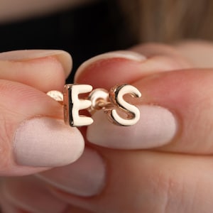 Peut inclure: Gros plan sur une main tenant une paire de boucles d'oreilles initiales en or. Les boucles d'oreilles sont en forme des lettres "E" et "S".