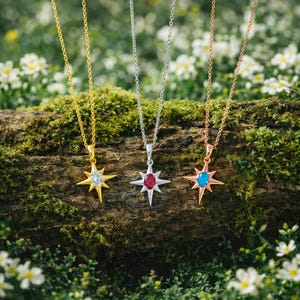 May include: Three starburst pendant necklaces in gold, silver, and rose gold. Each necklace features a different colored gemstone: clear, red, and blue. The necklaces are displayed on a moss-covered log with a blurred floral background.