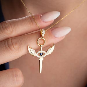 May include: Gold necklace with a pendant featuring a sword with wings and a blue evil eye. The pendant is adorned with clear cubic zirconia stones.