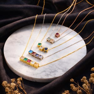 May include: A collection of gold and silver necklaces with square-shaped pendants. Each pendant features a different colored gemstone, including green, red, blue, and clear stones. The necklaces are displayed on a white marble surface against a black background.