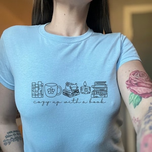 Può includere: T-shirt azzurra con un disegno a tratto nero di libri, una tazza, un gatto sui libri, una candela e altri libri. Il testo "cozy up with a book" è sotto i disegni.