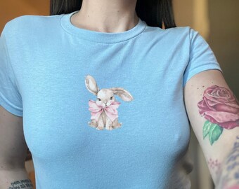 May include: A light blue t-shirt with a cartoon bunny wearing a pink bow. The bunny is in the center of the shirt.