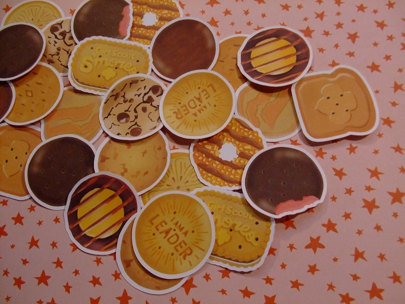 Op de afbeelding: Een verzameling Girl Scout cookie-stickers met verschillende smaken, waaronder Samoas, Tagalongs en Thin Mints. De stickers zijn gerangschikt op een roze achtergrond met oranje sterren. Sommige stickers bevatten de tekst "Ik ben een leider" en "Girl Scout Cookies".