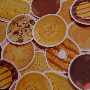 Op de afbeelding: Een verzameling Girl Scout cookie-stickers met verschillende smaken, waaronder Samoas, Thin Mints, Tagalongs en Do-si-dos. De stickers zijn rond en hebben kleurrijke illustraties van de koekjes.