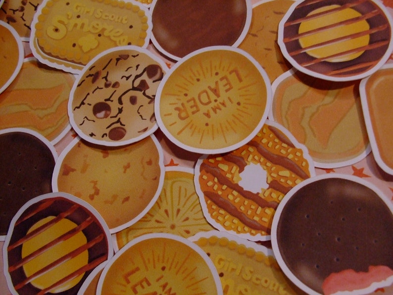 Op de afbeelding: Een verzameling Girl Scout cookie-stickers met verschillende smaken, waaronder Samoas, Thin Mints, Tagalongs en Do-si-dos. De stickers zijn rond en hebben kleurrijke illustraties van de koekjes.