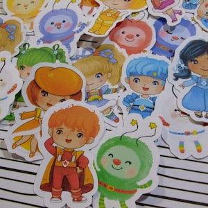 May include: A collection of colorful, die-cut stickers featuring cartoon characters with various hairstyles and outfits. The stickers include characters with blue, green, and orange hair, wearing costumes and dresses. Some stickers have star accents.