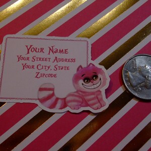 May include: Pink and white address label featuring a whimsical Cheshire Cat illustration.  The label includes fields for 'Your Name', 'Your Street Address', 'Your City, State', and 'Zipcode'. The Cheshire Cat is depicted in a playful, cartoonish style with pink and white stripes. The background of the label is a soft pink, and the text is a darker pink.  The overall style is reminiscent of Alice in Wonderland.