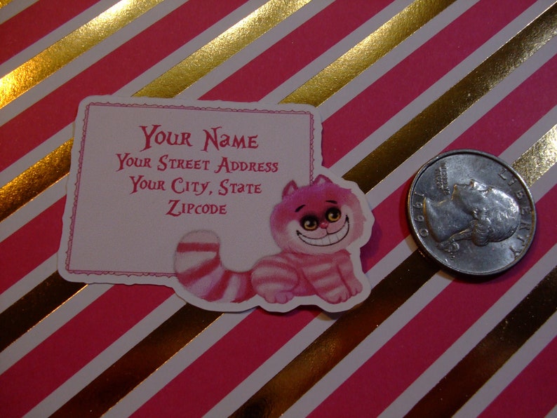 May include: Pink and white address label featuring a whimsical Cheshire Cat illustration.  The label includes fields for 'Your Name', 'Your Street Address', 'Your City, State', and 'Zipcode'. The Cheshire Cat is depicted in a playful, cartoonish style with pink and white stripes. The background of the label is a soft pink, and the text is a darker pink.  The overall style is reminiscent of Alice in Wonderland.