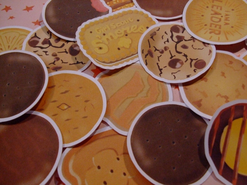 Op de afbeelding: Een verzameling ronde, kleurrijke en gepatroneerde koekjestickers. De stickers hebben verschillende ontwerpen, waaronder een Girl Scout S'mores-koekje, een chocoladekoekje en een pindakaaskoekje.