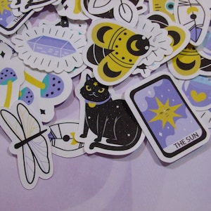 May include: A collection of colorful stickers featuring various designs, including a black cat, a bee, mushrooms, a dragonfly, a crystal, and a tarot card with the text "THE SUN".