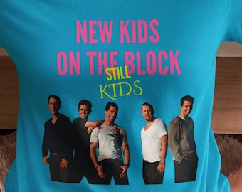 Könnte beinhalten: Ein türkisfarbenes T-Shirt mit dem Text "NEW KIDS ON THE BLOCK STILL KIDS" in Pink und Gelb. Das Shirt zeigt ein Gruppenfoto von fünf Männern. Das T-Shirt hat einen Rundhalsausschnitt.