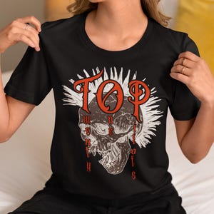 Könnte beinhalten: Schwarzes T-Shirt mit einem Totenkopf-Motiv und dem roten Text "TOP" und "UNITY OF SOULS". Der Totenkopf ist braun und weiß, mit einem weißen Burst-Design dahinter. Das T-Shirt hat kurze Ärmel und einen Rundhalsausschnitt.