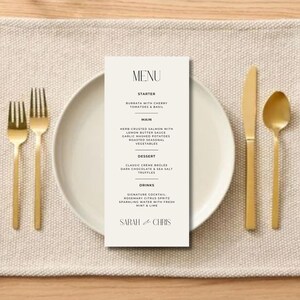 May include: A white menu card with black text listing the menu items for a wedding reception. The menu includes a starter, main course, dessert, and drinks. The card is on a beige placemat with gold silverware and a white plate.