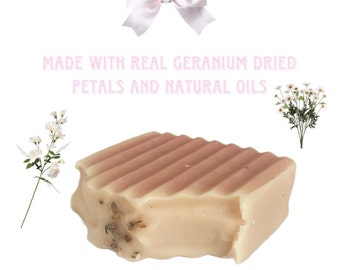 May include: A bar of handmade soap with a light beige color and a textured top. The soap contains dried petals and natural oils. The text "MADE WITH REAL GERANIUM DRIED PETALS AND NATURAL OILS" is displayed in pink.