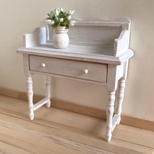 May include: A miniature white desk with a single drawer and turned legs. A small white vase with green and white flowers sits on the desk's surface. The desk has a distressed, vintage appearance, suitable for dollhouses or decorative displays.