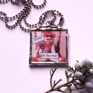 May include: A silver-toned necklace with a square pendant featuring a sock monkey wearing sunglasses and a red hat. The pendant has the text "Always get the red!" on it. The background is a light pink.