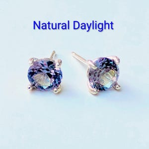 May include: A pair of round-cut stud earrings featuring faceted, colour-shifting purple and blue gemstones set in four-prong metal settings. The text 'Natural Daylight' is positioned above the earrings, indicating the lighting conditions for the display.