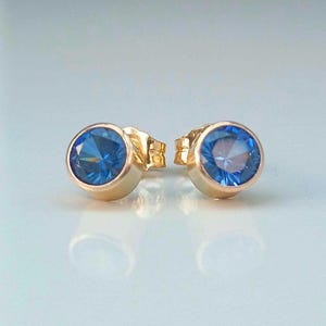 Könnte beinhalten: Ein Paar goldene Ohrstecker mit runden, facettierten blauen Edelsteinen. Die Ohrringe haben ein schlichtes, klassisches Design mit einer Lünettenfassung. Die Edelsteine sind in einem leuchtenden Blau gehalten.