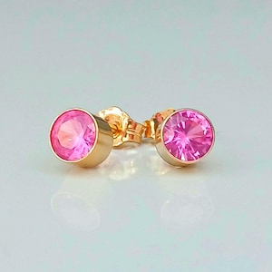 May include: A pair of gold stud earrings featuring round, bright pink gemstones. The earrings have a classic design with a bezel setting. The gemstones are a vibrant pink colour, and the gold setting adds a touch of elegance.