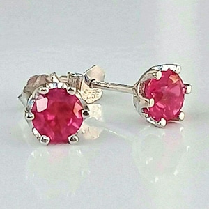 Puede incluir: Un par de pendientes de botón con piedras redondas facetadas de color rosa vibrante engastadas en garras de metal plateado. Cada pendiente tiene un cierre de presión y un sello 925 visible en el metal, indicando plata de ley en el engaste.