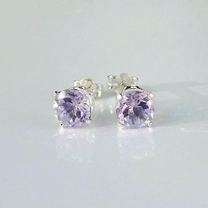 May include: A pair of round-cut, light purple amethyst stud earrings set in polished silver metal. Each gemstone features a faceted surface that reflects light, secured by four prongs on a silver post with matching silver butterfly earring backs.