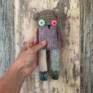 Könnte beinhalten: Ein gestrickter Spielzeugbär mit einem lila Körper, einem braunen Kopf und mehrfarbigen Armen und Beinen. Der Bär hat zwei große Knöpfe für Augen, einen grünen und einen rosa.