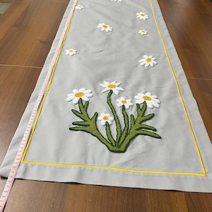 Op de afbeelding: Een grijze tafelrunner met een gele rand en geborduurde witte madeliefjes met groene stelen. De runner ligt op een houten tafel.