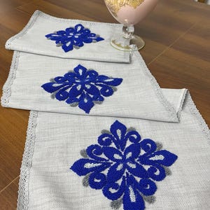 May include: Three gray table runners with blue and gray embroidered floral designs. The runners have a white lace trim.