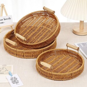 May include: Three round woven wicker trays with light brown finish and smooth wooden handles. The trays feature a circular base and vertical sides, with two handles attached to the rim of each tray for carrying. The set includes trays of varying sizes.