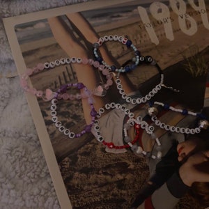 Könnte beinhalten: Eine Sammlung von Perlenarmbändern mit verschiedenen farbigen Perlen und weißen Buchstabenperlen, die Sätze wie "Lover", "Speak Now", "It's So Hard", "Rureadyforit", "Anyway" und "Mirrorball" buchstabieren.