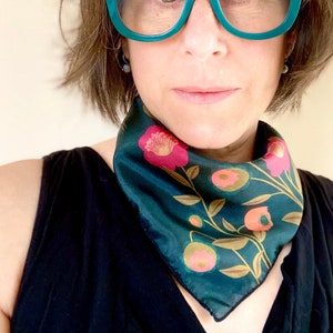 May include: A woman wearing a teal green bandana with a floral pattern and teal green cat-eye glasses. The bandana has a dark green background with pink and yellow flowers.