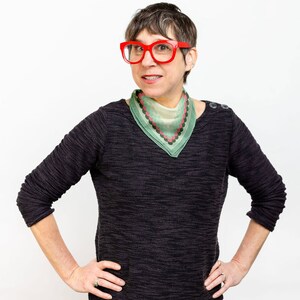 May include: A woman with short brown hair wearing red glasses and a green and pink patterned bandana. She is wearing a black sweater and black and white patterned pants.