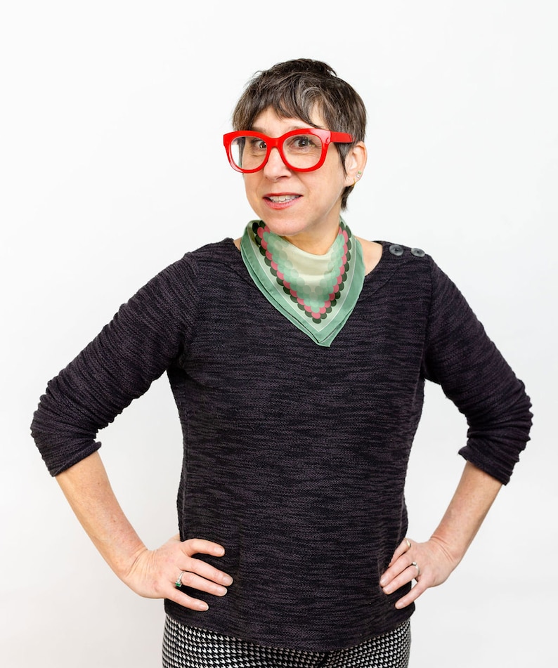 May include: A woman with short brown hair wearing red glasses and a green and pink patterned bandana. She is wearing a black sweater and black and white patterned pants.