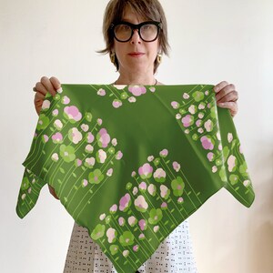 May include: A green silk scarf with a pink and green floral pattern. The scarf is folded in half and held by a person wearing glasses.