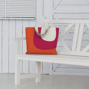 May include: A white canvas tote bag with orange and pink geometric shapes. The bag is sitting on a white wooden bench.