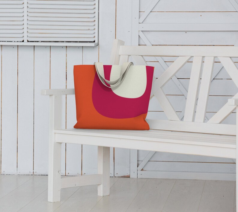 May include: A white canvas tote bag with orange and pink geometric shapes. The bag is sitting on a white wooden bench.