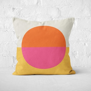 Peut inclure: Un coussin décoratif avec un motif géométrique. La moitié supérieure est crème, avec un grand cercle orange au centre. La moitié inférieure est jaune, avec un cercle rose. Le coussin est sur une surface blanche.