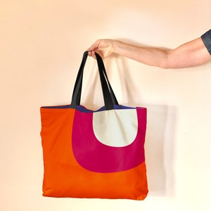 May include: A large orange tote bag with a pink and white geometric design. The bag has black straps and a blue lining.