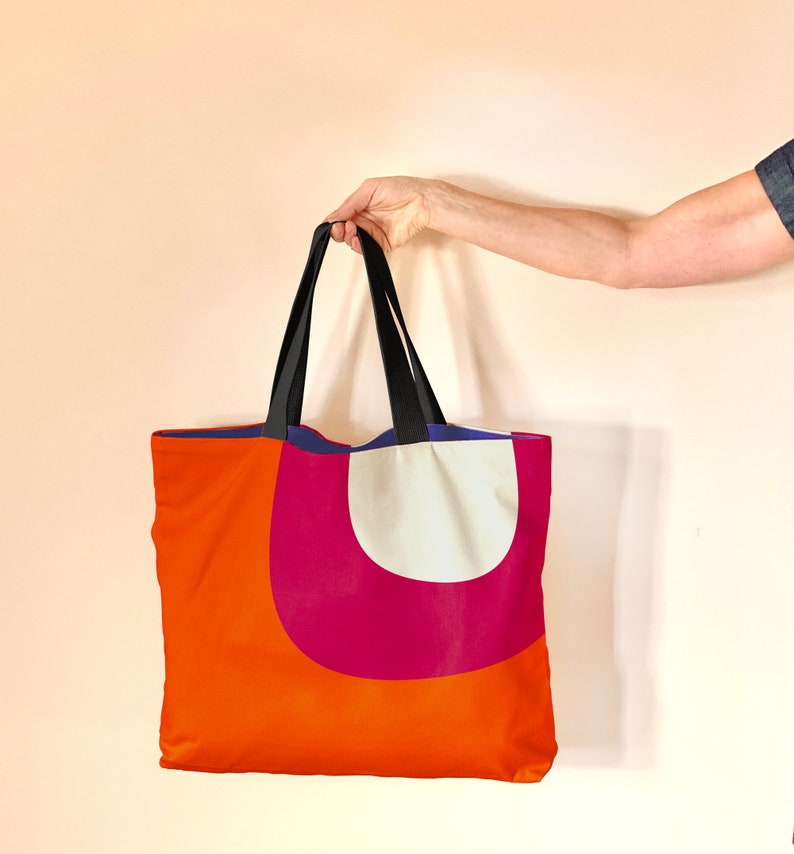 May include: A large orange tote bag with a pink and white geometric design. The bag has black straps and a blue lining.