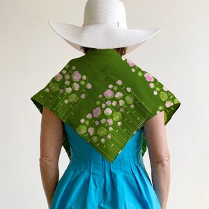 May include: A woman wearing a white sun hat and a green scarf with pink and green floral print. The scarf is tied around her neck and drapes down her back. She is wearing a bright blue dress.